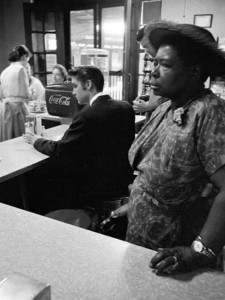 Elvis sits to eat at a segregated lunch counter while an elderly black woman stands, waiting for food to take away. she's not allowed to sit there.