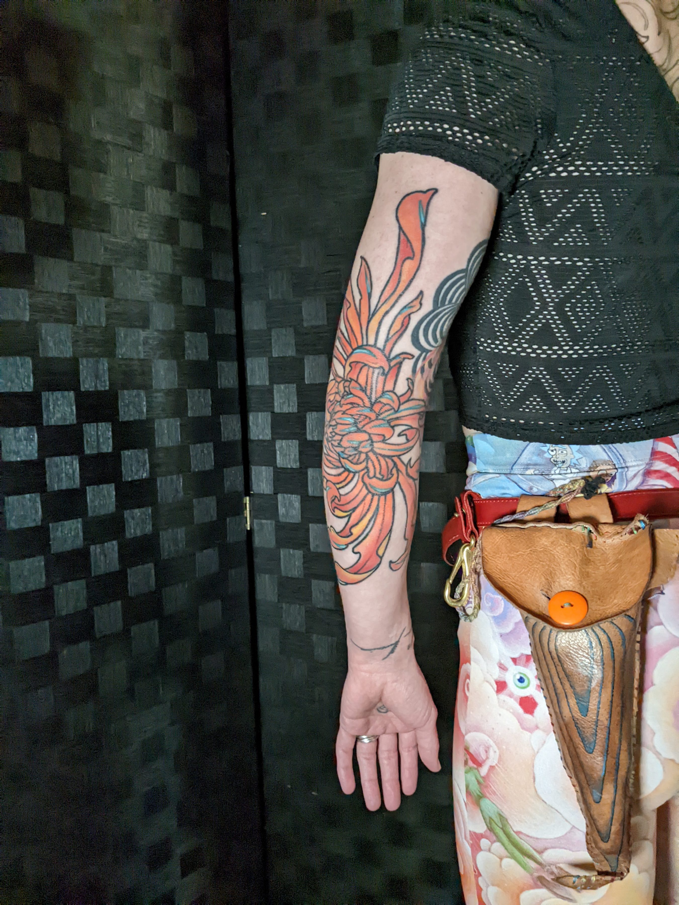 a tattoo of a bright orange, large chrysanthemum on an elbow the person is wearing a matching hip sack, jeans, and a black shirt
